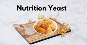 bowl of golden-brown, popcorn-coated balls sits on a wooden cutting board. A small bowl of nutrition yeast is next to the cutting board, and a few scattered popcorn kernels are around the bowl.